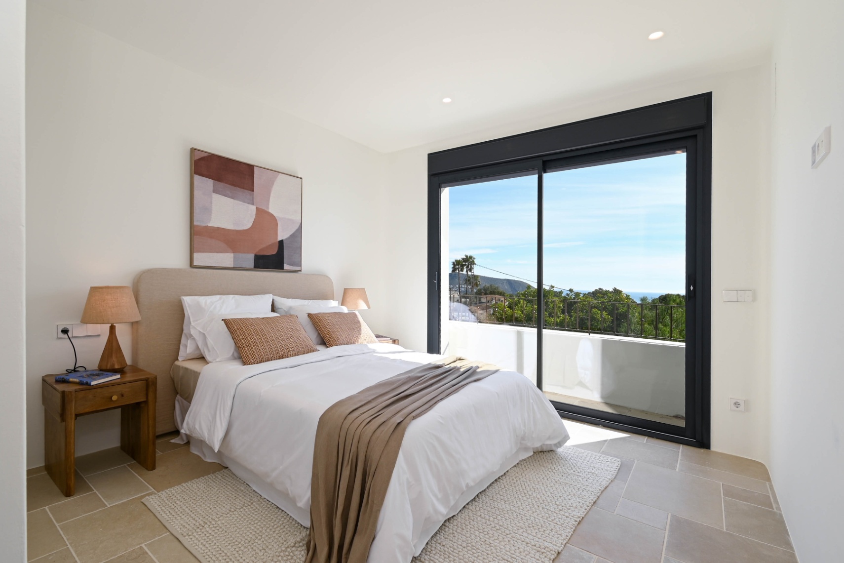 Dormitorio luminoso con suelos de piedra natural y grandes ventanales correderos que dan acceso a una terraza privada con vistas despejadas al entorno mediterráneo.