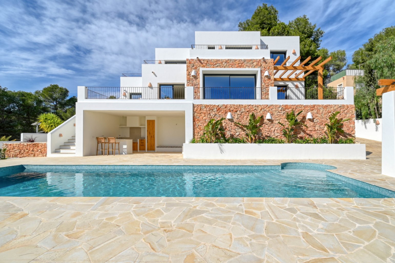 Exterior de la villa con piscina de mosaico azul, terraza de piedra natural y fachada de tres niveles que integra muros de piedra, cocina exterior y una pérgola de madera.