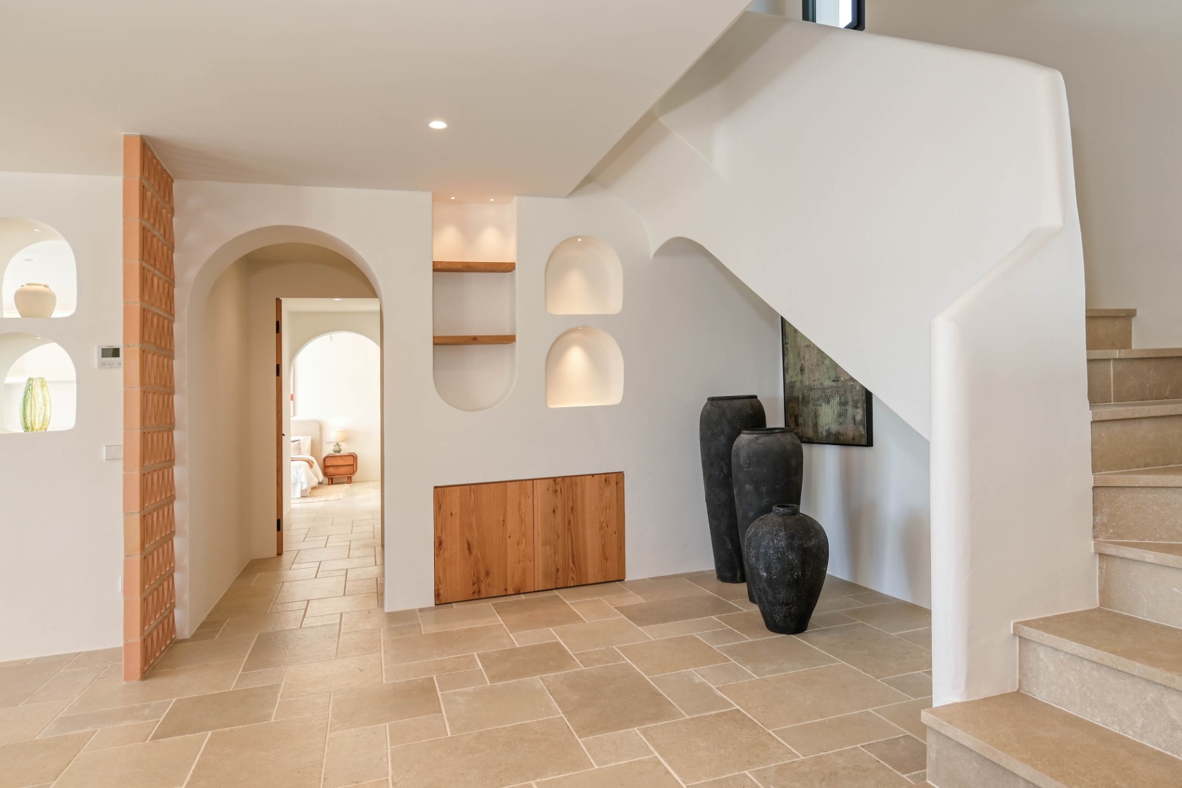 Vestíbulo de diseño mediterráneo con escalera de piedra, hornacinas iluminadas y armarios de madera integrados. El suelo de piedra natural conecta con elegantes arcos.