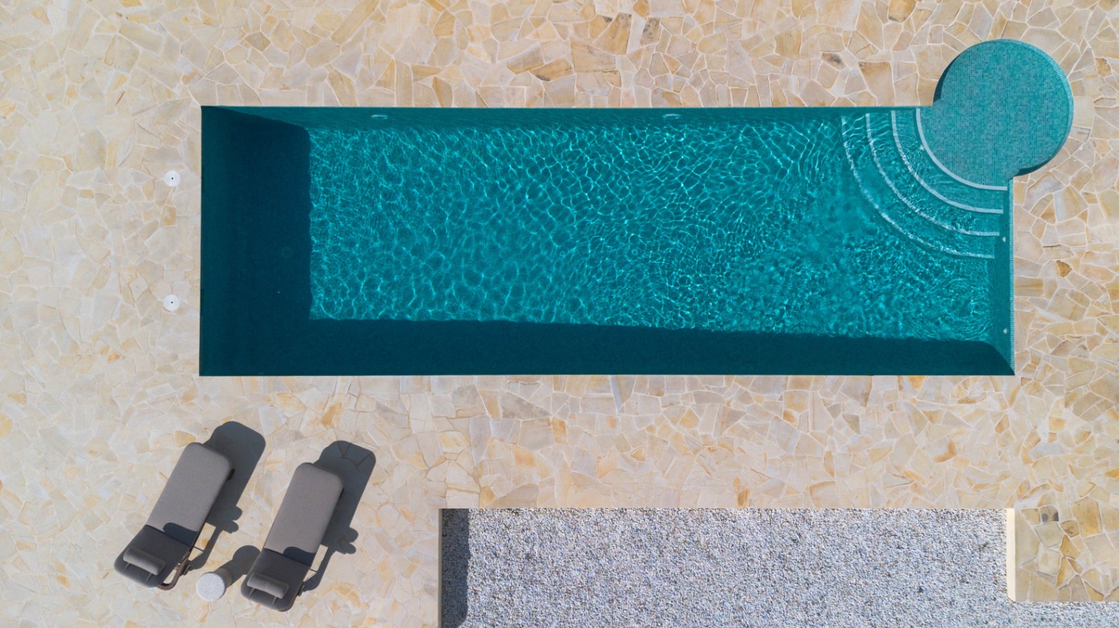 Vista cenital de la piscina de 13 metros con gresite turquesa, rodeada por una terraza de piedra natural irregular y zona de descanso con tumbonas.