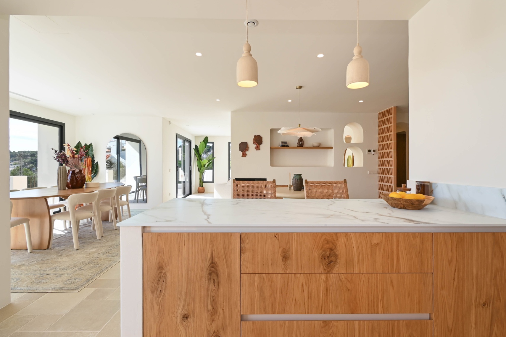 Vista desde la isla de cocina con encimera de piedra veteada hacia el comedor, resaltando los nichos decorativos, el mobiliario de madera y la abundante luz natural.