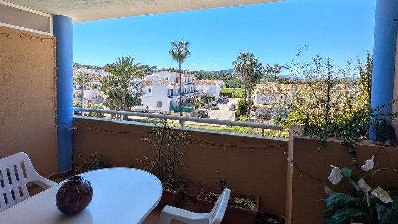 Balcón con mesa y silla, ofreciendo vistas despejadas a una zona residencial con casas blancas, palmeras y colinas verdes bajo un cielo azul brillante. Balcón con mesa y silla, ofreciendo vistas despejadas a una zona residencial con casas blancas, palmeras y colinas verdes bajo un cielo azul brillante.
