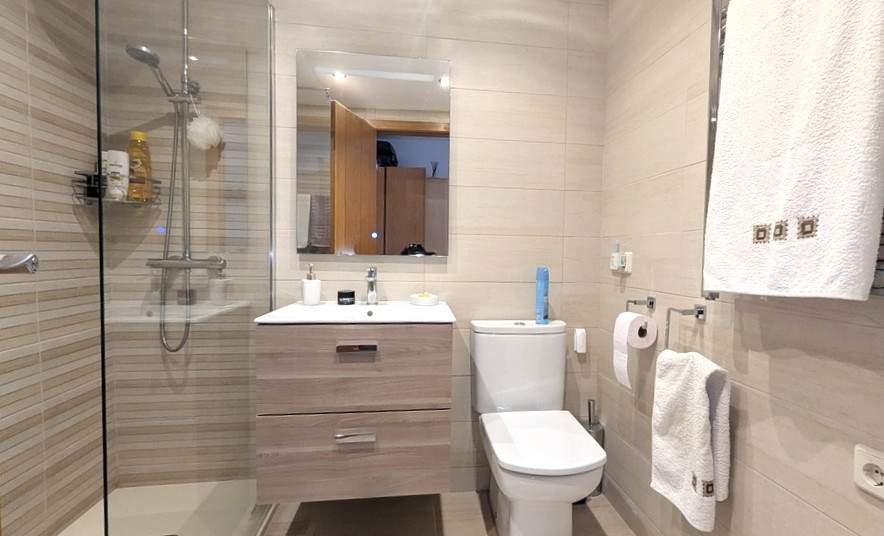 Baño moderno con ducha de obra acristalada, mueble lavabo suspendido en acabado madera clara y revestimiento cerámico de tonos neutros.