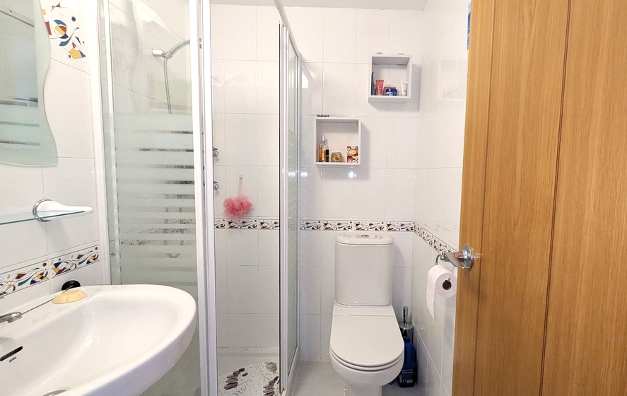 Cuarto de baño con ducha, paredes de azulejos blancos con cenefa decorativa, mampara de cristal translúcido y estantes auxiliares de pared.