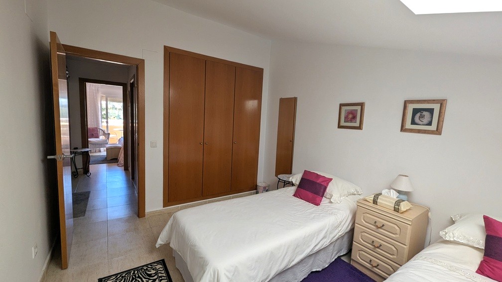 Dormitorio con dos camas individuales, armarios empotrados de madera y suelos cerámicos. La puerta abierta conecta con el pasillo y una estancia con vistas exteriores.