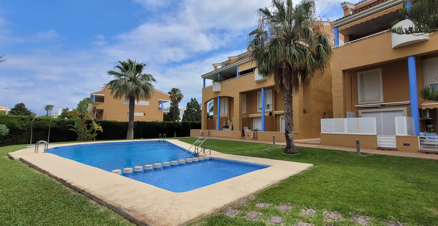 La atractiva piscina comunitaria, con singulares escalones en su parte menos profunda, está rodeada de césped cuidado y edificios residenciales de tonos cálidos con acentos azules y palmeras. La atractiva piscina comunitaria, con singulares escalones en su parte menos profunda, está rodeada de césped cuidado y edificios residenciales de tonos cálidos con acentos azules y palmeras.