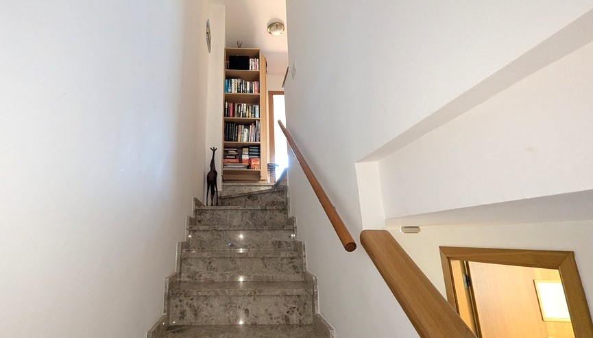 La elegante escalera interior de mármol pulido con iluminación integrada y barandilla de madera conduce al nivel superior con una estantería llena de libros. La elegante escalera interior de mármol pulido con iluminación integrada y barandilla de madera conduce al nivel superior con una estantería llena de libros.