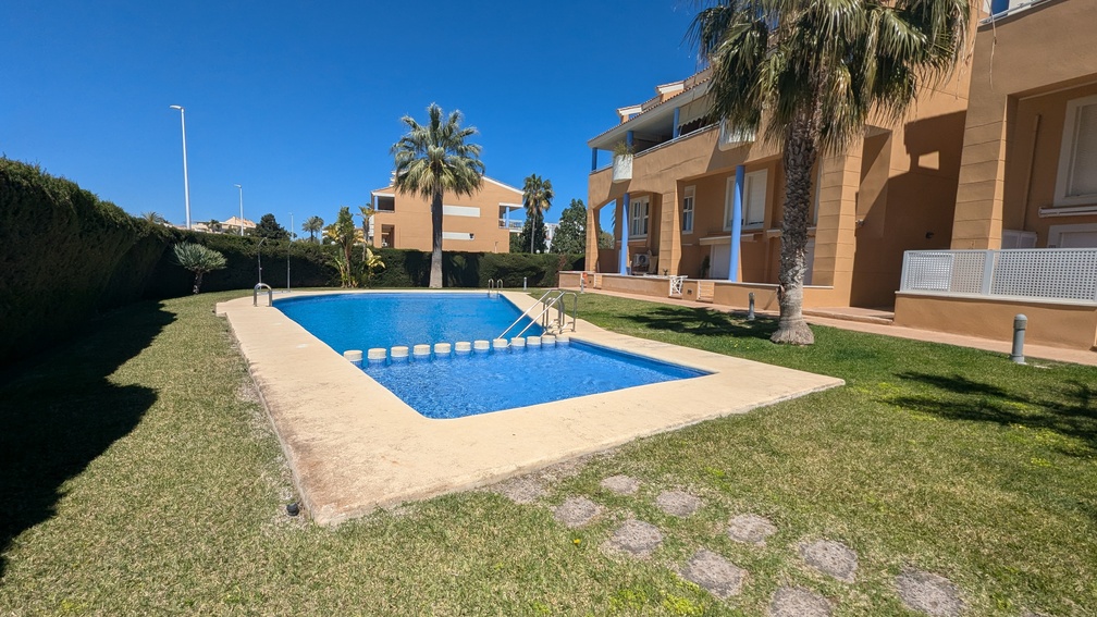 Piscina comunitaria con zona infantil integrada, rodeada de césped cuidado, palmeras y la fachada de tonos ocres del complejo residencial en Jávea.