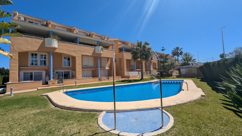 Piscina comunitaria de forma arriñonada rodeada de césped y jardines mediterráneos, con la fachada de color terracota y pilares azules del edificio al fondo.
