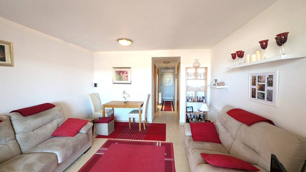 Salón-comedor con sofás beige, detalles rojos y mesa de madera. El espacio conecta con un pasillo central, con suelos de gres claro y paredes blancas.