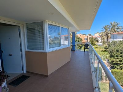 Amplia terraza privada con suelo de baldosas marrones, barandilla de cristal y cerramiento parcial. Ofrece vistas a palmeras y al paisaje residencial local.