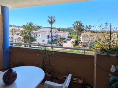 Balcón con mesa y silla, ofreciendo vistas despejadas a una zona residencial con casas blancas, palmeras y colinas verdes bajo un cielo azul brillante.