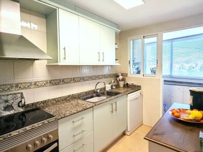 Cocina totalmente equipada con armarios claros, encimeras tipo granito, placa y horno integrados, y ventana a una zona de lavadero adyacente.