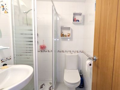 El moderno cuarto de ducha presenta paredes alicatadas en blanco con un borde decorativo, una mampara de cristal esmerilado, un lavabo suspendido y un inodoro. Dos estantes cuadrados ofrecen almacenamiento práctico.