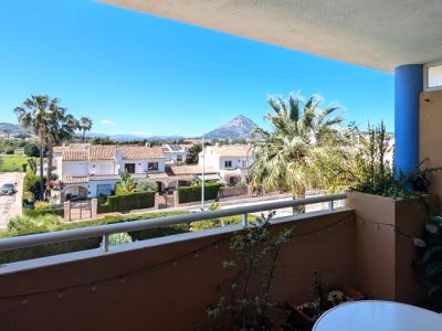 Terraza abierta con barandilla blanca y plantas, ofreciendo vistas panorámicas a residencias locales, zonas verdes y la distintiva montaña del Montgó bajo un cielo despejado.