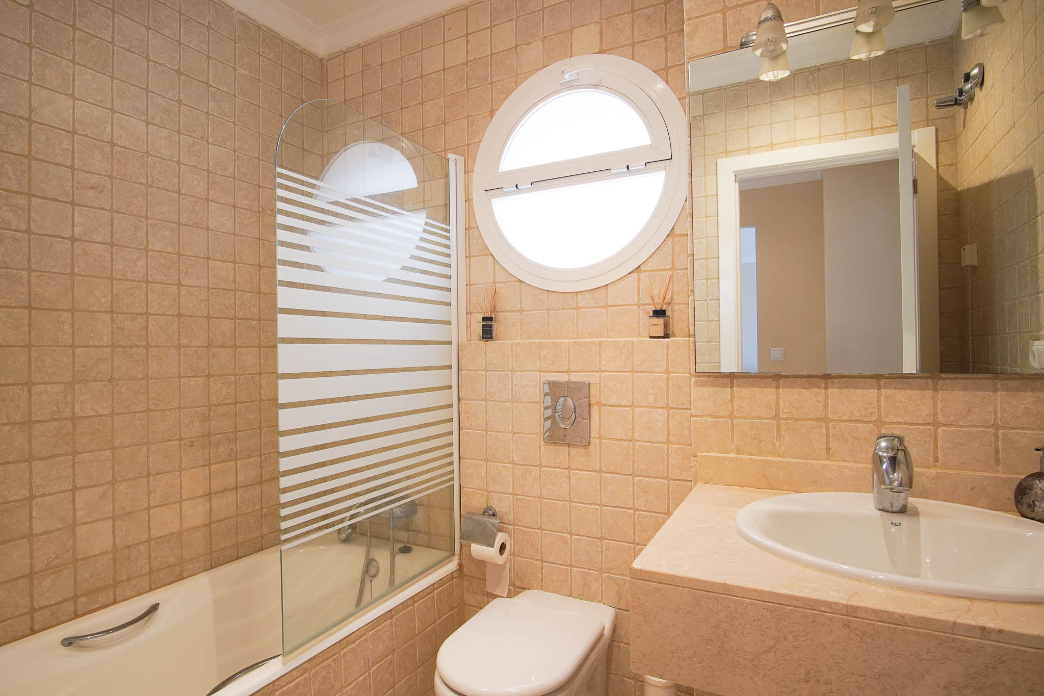 Baño con encimera de mármol, bañera con mampara de cristal serigrafiado y una característica ventana circular que aporta luz natural.
