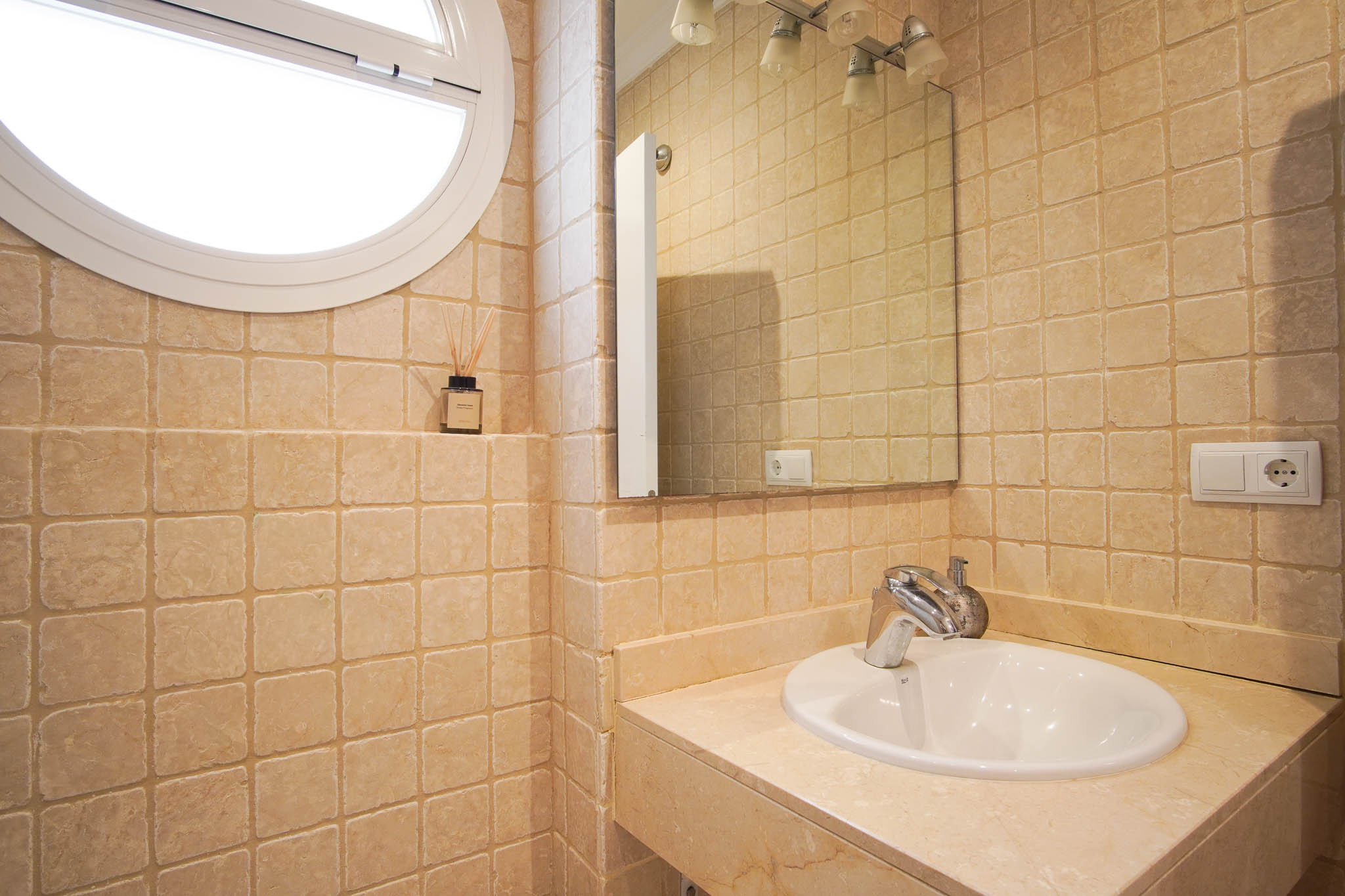 Baño con revestimiento de mármol abujardado, lavabo encastrado en encimera de piedra y una característica ventana circular que aporta luz natural.