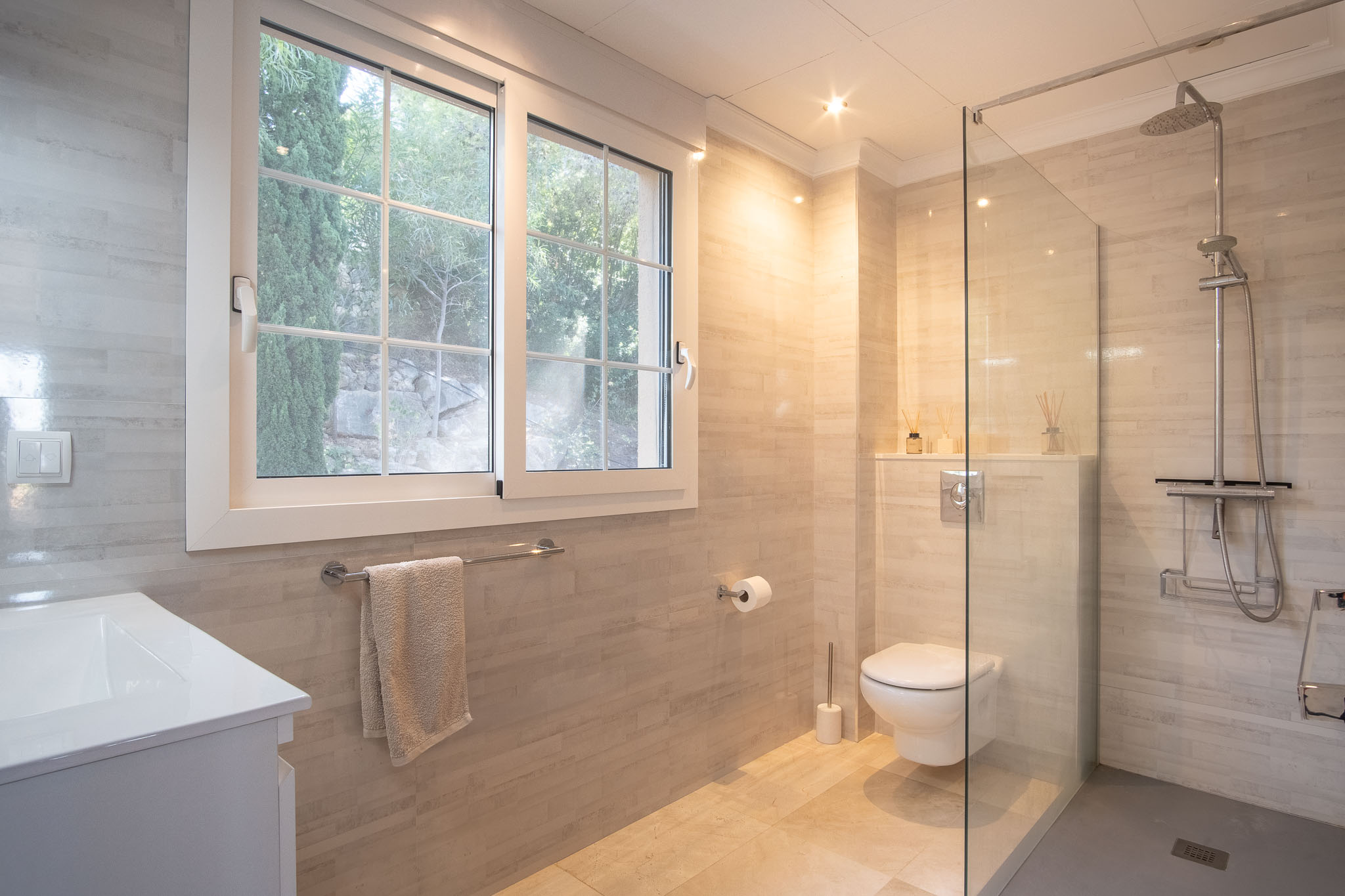 Baño moderno con ducha a ras de suelo, mampara de cristal y revestimiento cerámico en tonos crema. Incluye sanitarios suspendidos y una ventana que aporta luz natural y vistas al jardín.