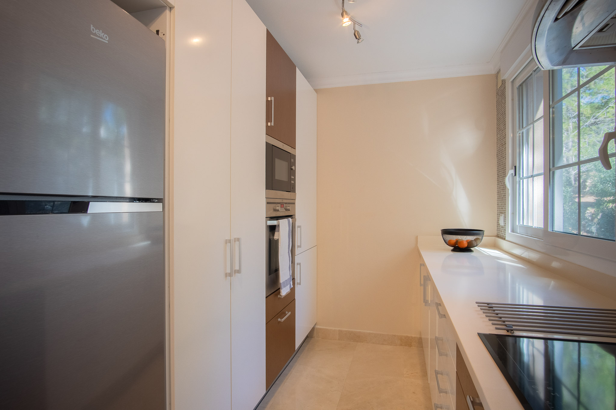 Cocina moderna con armarios en blanco y madera, electrodomésticos de acero inoxidable integrados y encimera blanca. Una ventana lateral aporta luz natural sobre la zona de cocción.