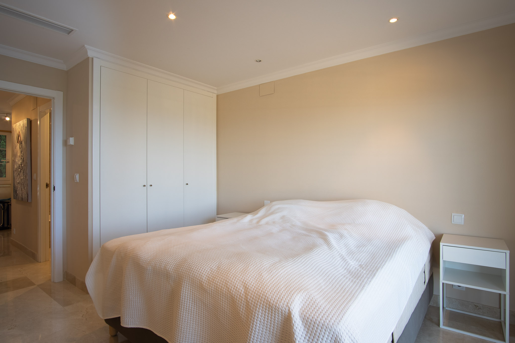 Dormitorio con suelos de mármol, amplios armarios empotrados lacados en blanco e iluminación mediante focos empotrados en el techo, integrando un diseño sobrio y funcional.