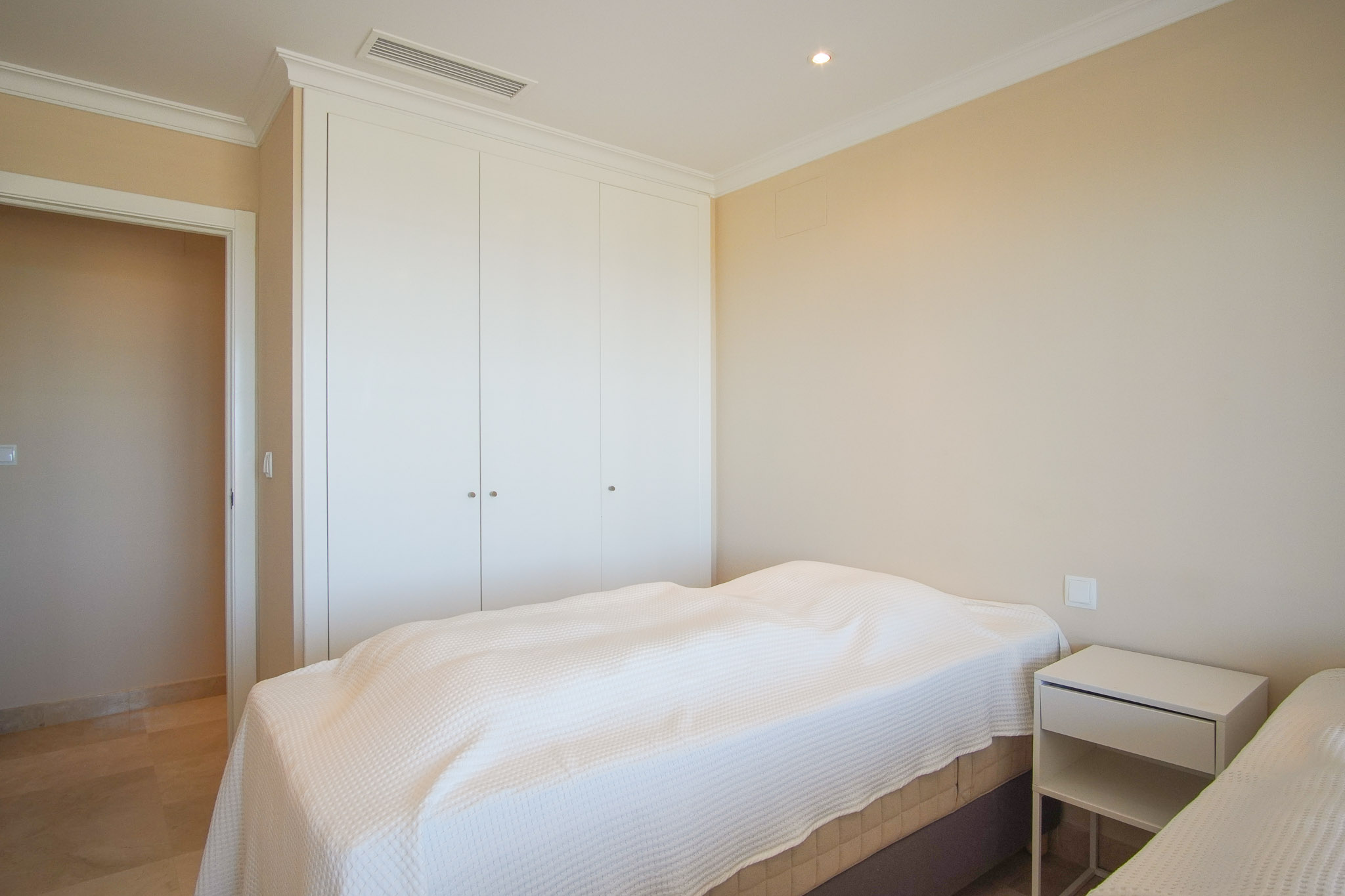 Dormitorio luminoso con armarios empotrados blancos, suelos de mármol y climatización integrada. Paredes en tonos crema y molduras clásicas en el techo.