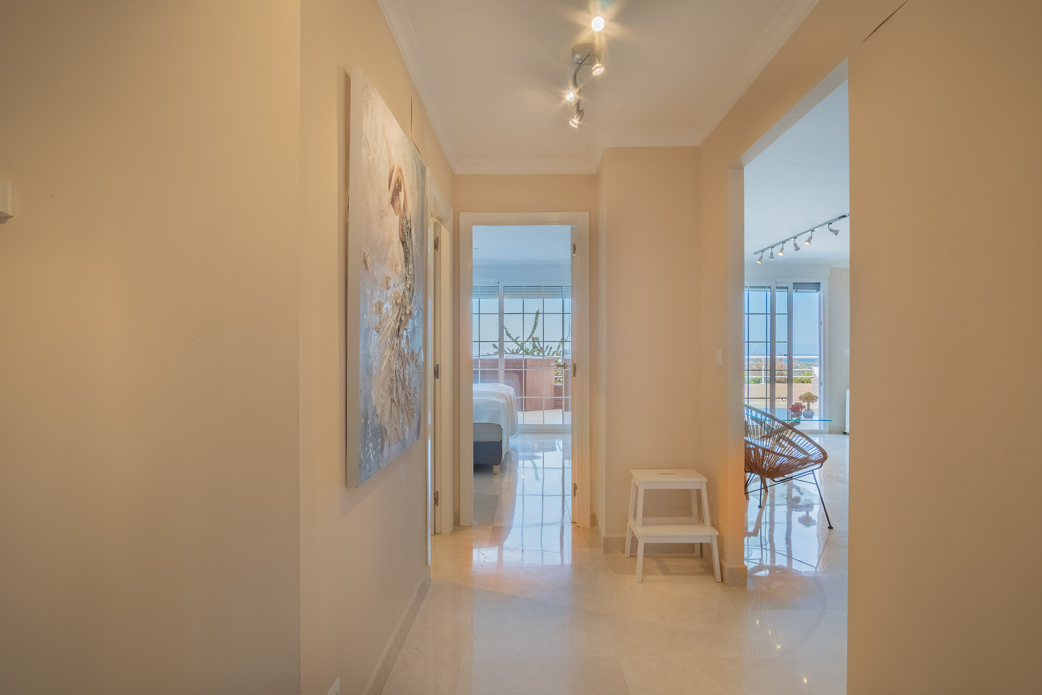 Pasillo con suelos de mármol y focos direccionales, que conecta las estancias principales y ofrece una vista directa hacia un dormitorio con acceso a la terraza.