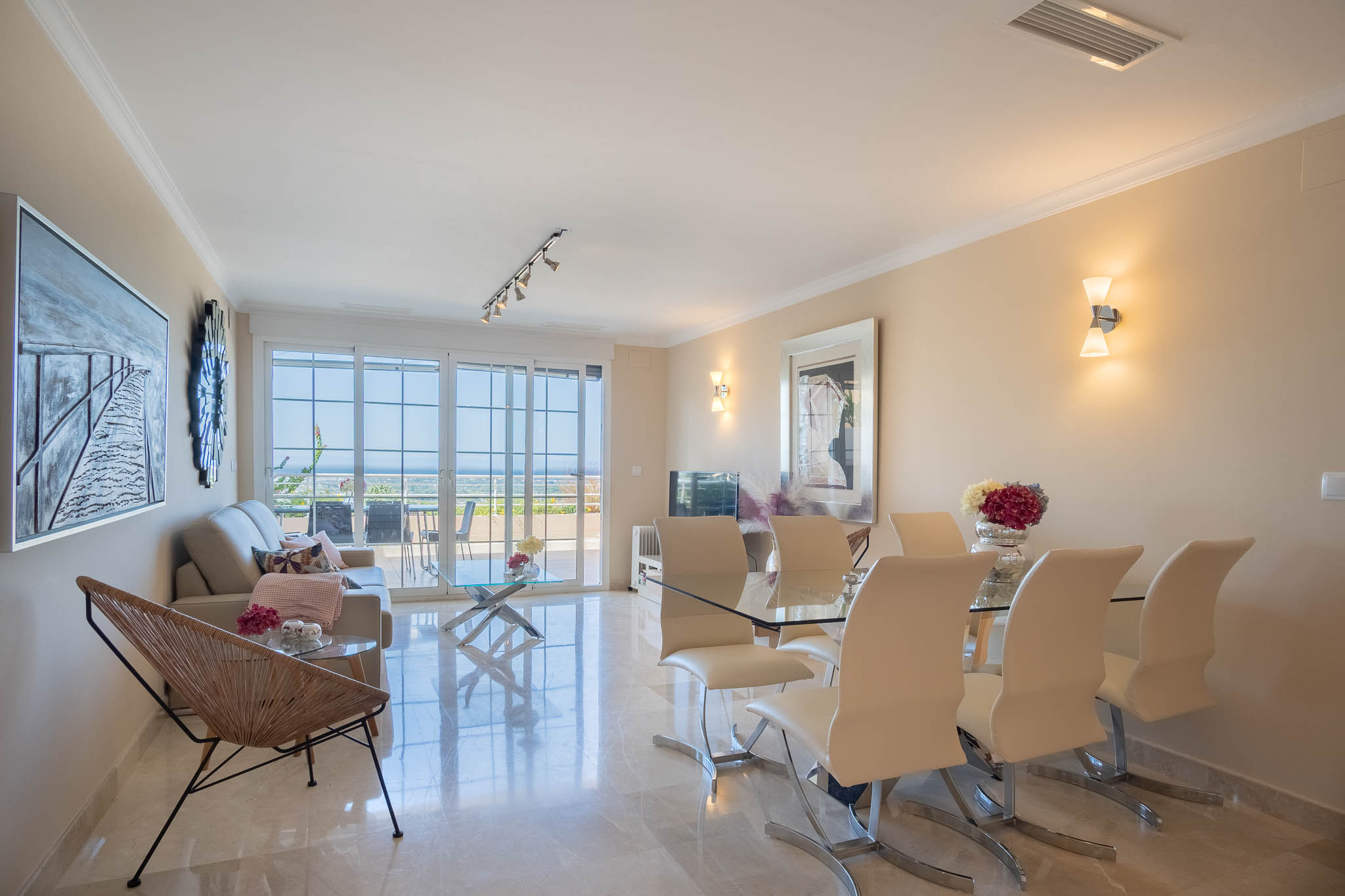 Salón-comedor de planta abierta con suelos de mármol pulido, mobiliario moderno de cristal y acceso directo a la terraza con vistas al mar a través de grandes ventanales.