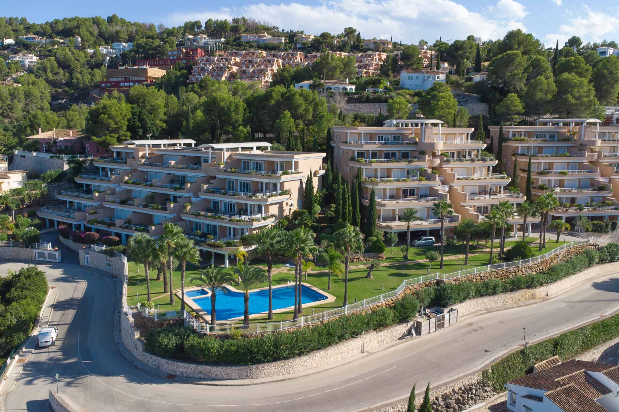 Vista aérea del complejo Las Mimosas en La Sella, mostrando su arquitectura aterrazada, la piscina comunitaria rodeada de palmeras y las extensas zonas verdes.