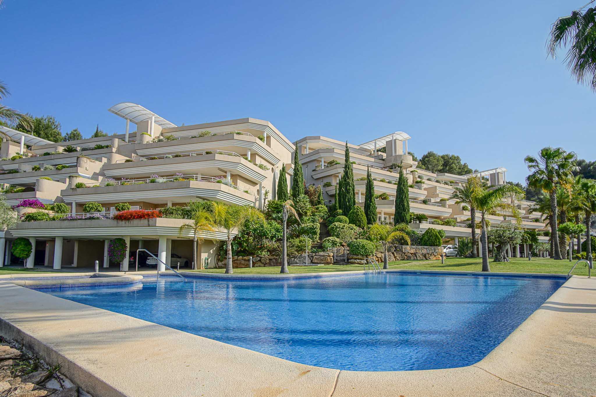 Vista exterior del complejo Las Mimosas, destacando su piscina comunitaria y la arquitectura escalonada con amplias terrazas rodeadas de vegetación mediterránea.