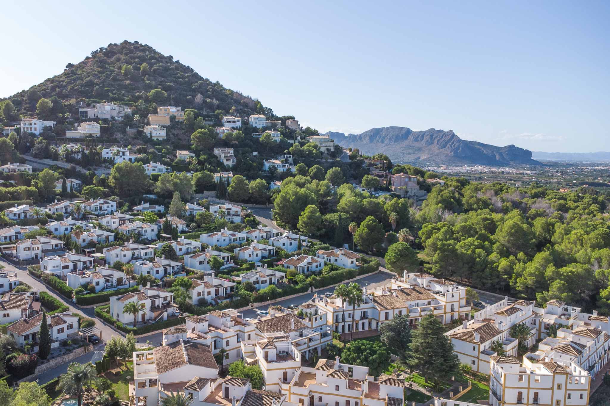 Vista panorámica elevada del complejo residencial La Sella, destacando su arquitectura mediterránea entre frondosos pinos y con las montañas al fondo.