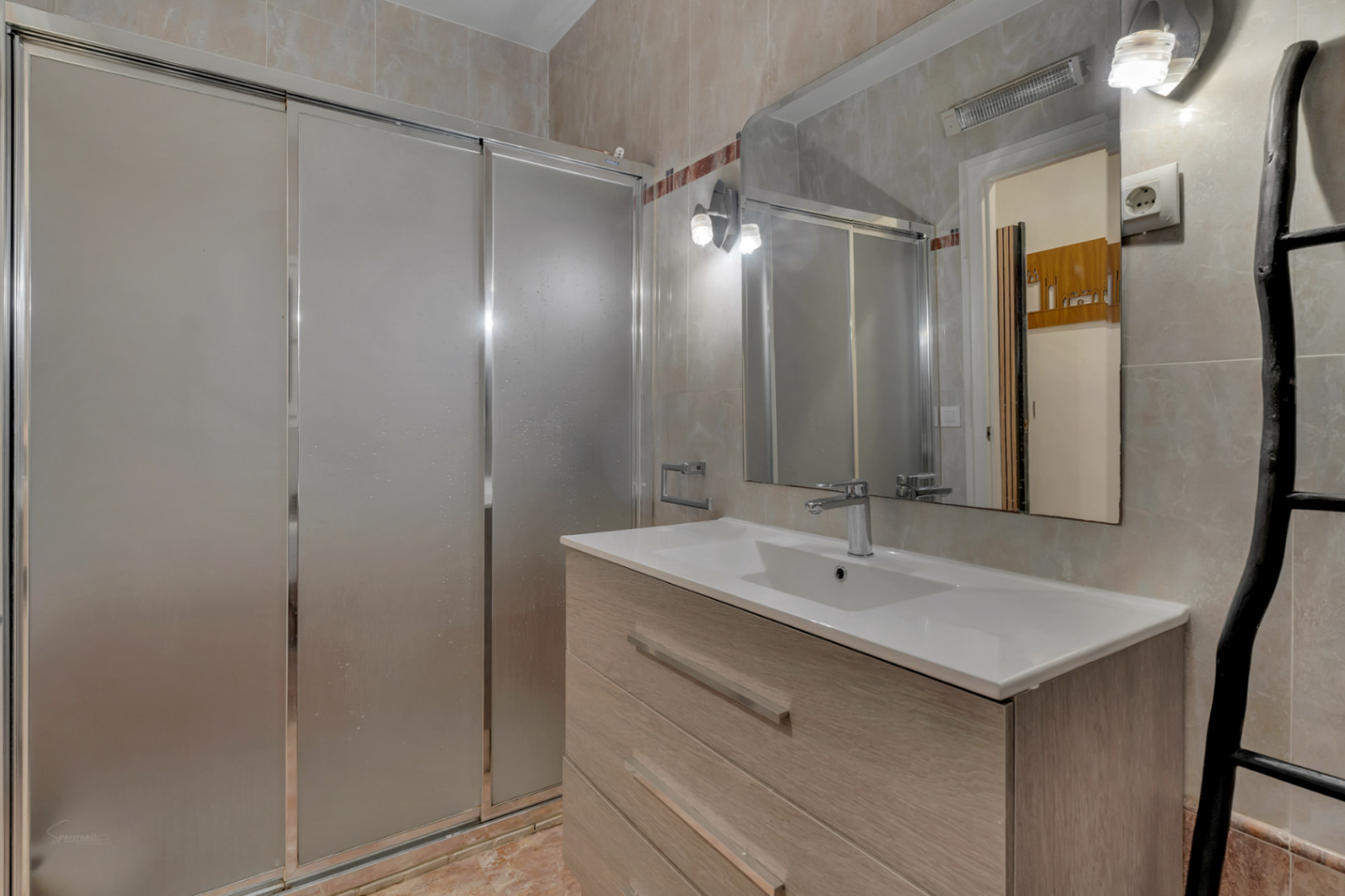 Detalle del baño con ducha corredera, lavabo moderno con mueble de madera clara y espejo con iluminación.