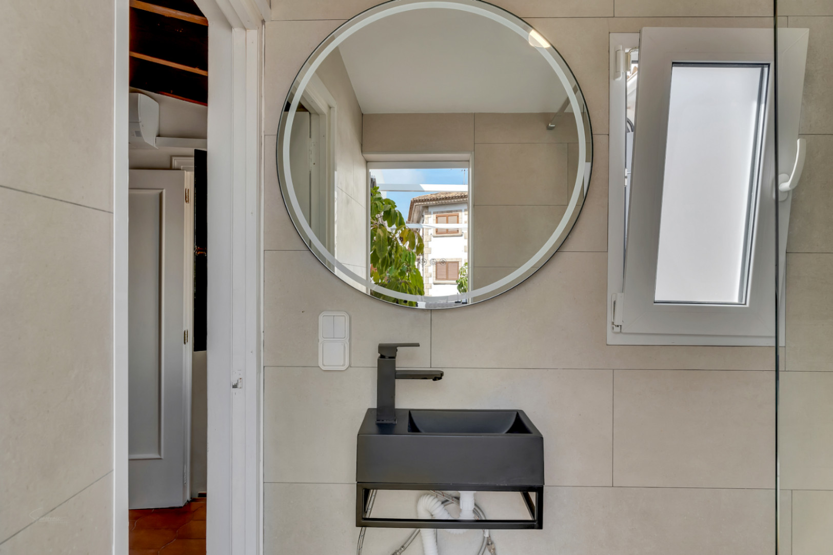 Detalle del baño con lavabo negro moderno, espejo circular iluminado y ventana abatible.