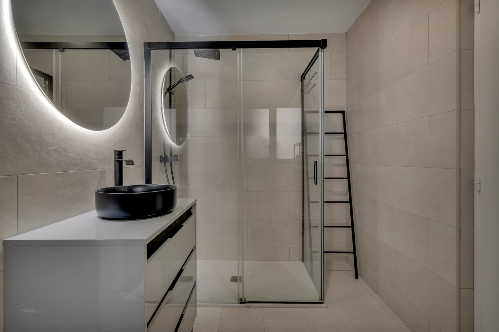 Detalle del baño moderno con lavabo negro sobre encimera blanca y ducha con mampara de cristal.