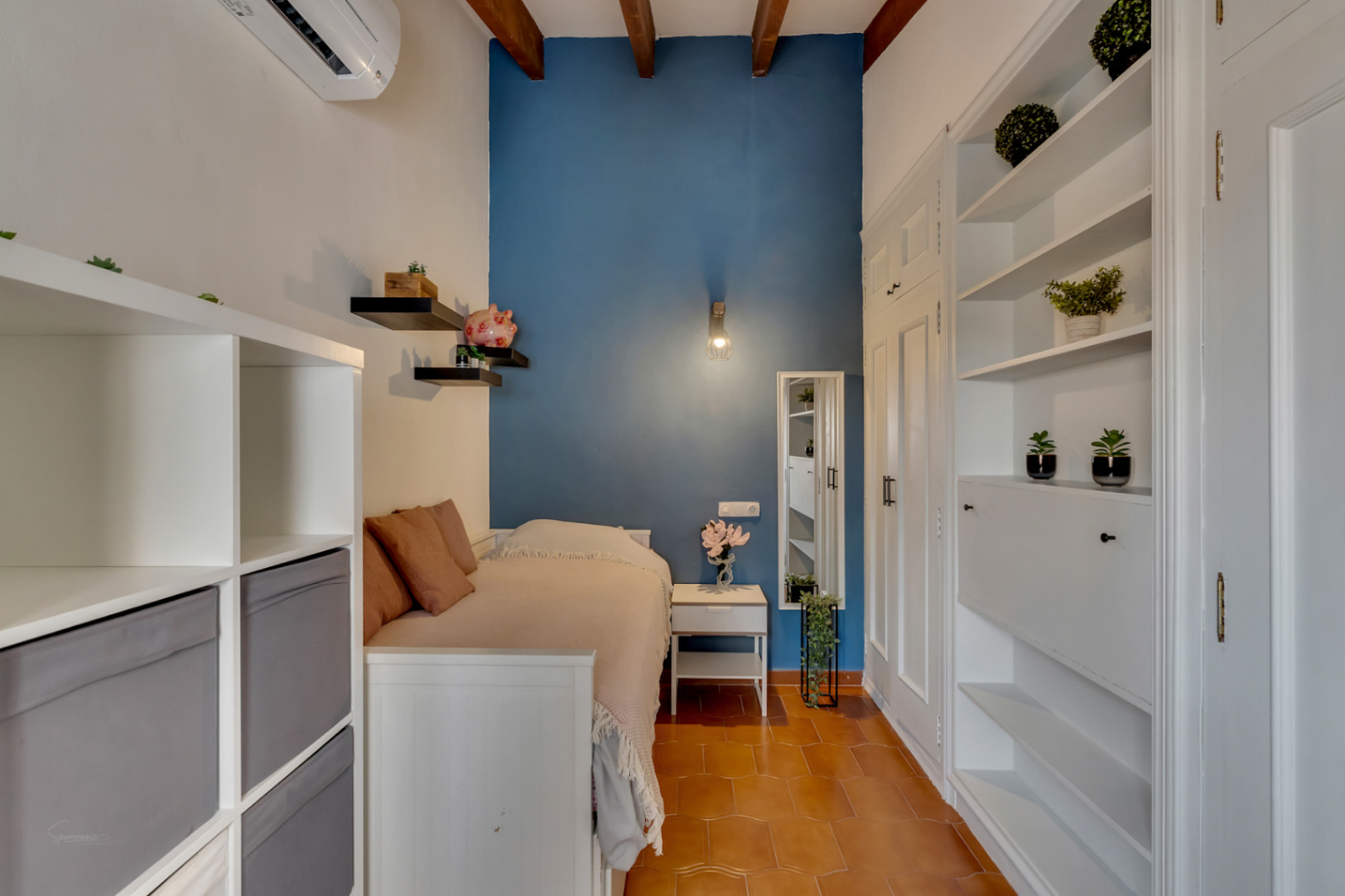 Dormitorio con pared azul, estanterías blancas y suelo de terracota. Vigas de madera en el techo.