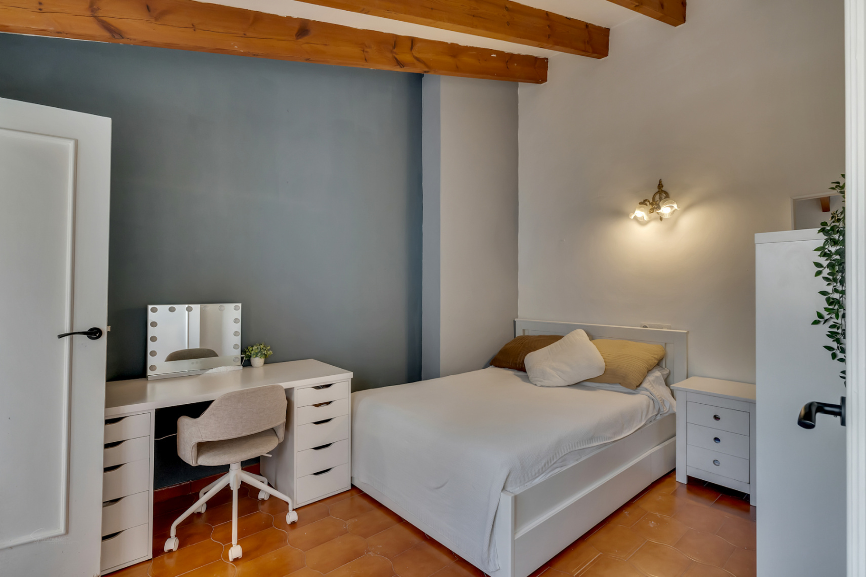 Dormitorio con suelo de terracota, pared azul y escritorio blanco con espejo iluminado.