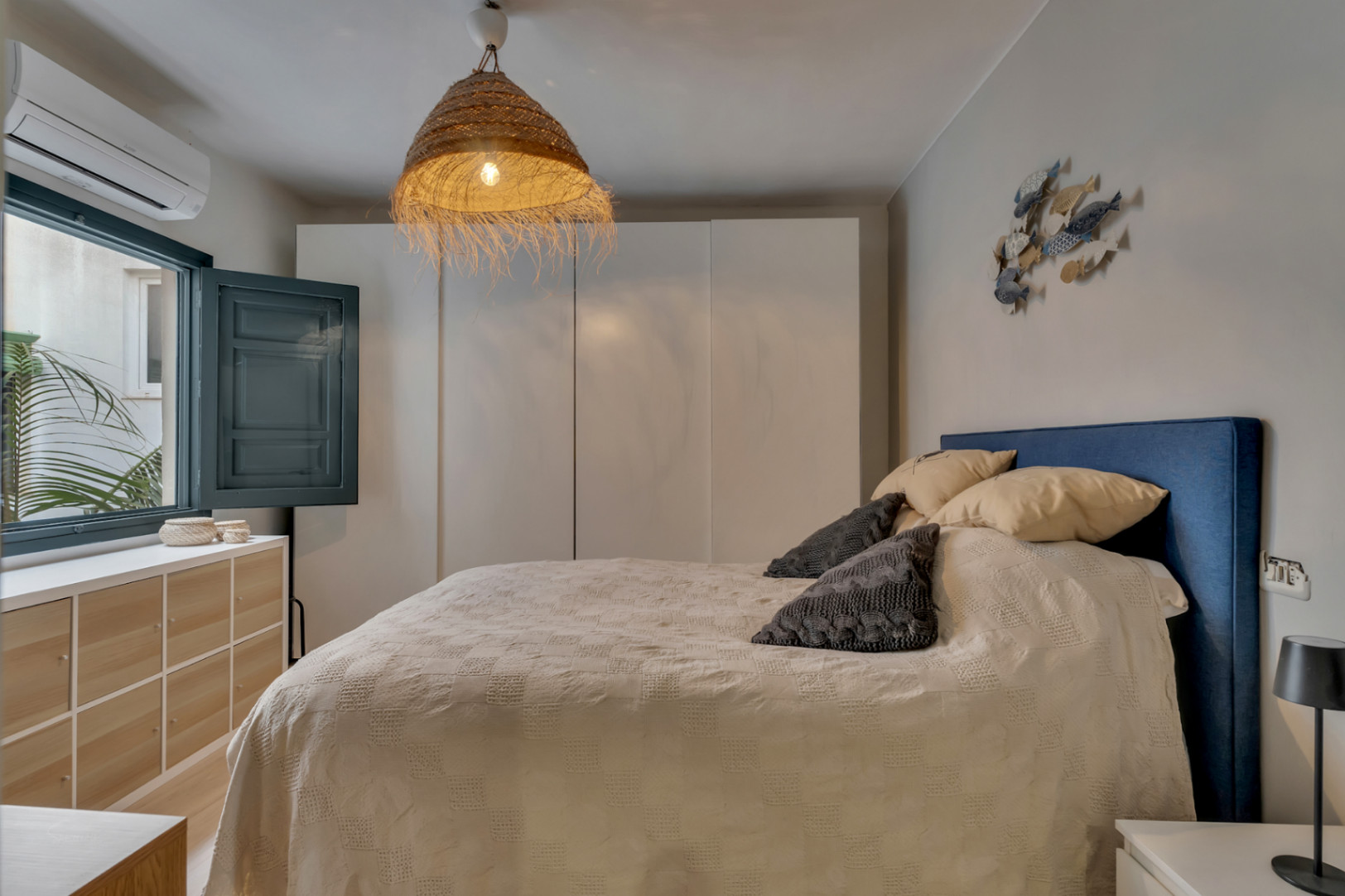 Dormitorio luminoso con cabecero azul, lámpara de mimbre y ventana con contraventanas.