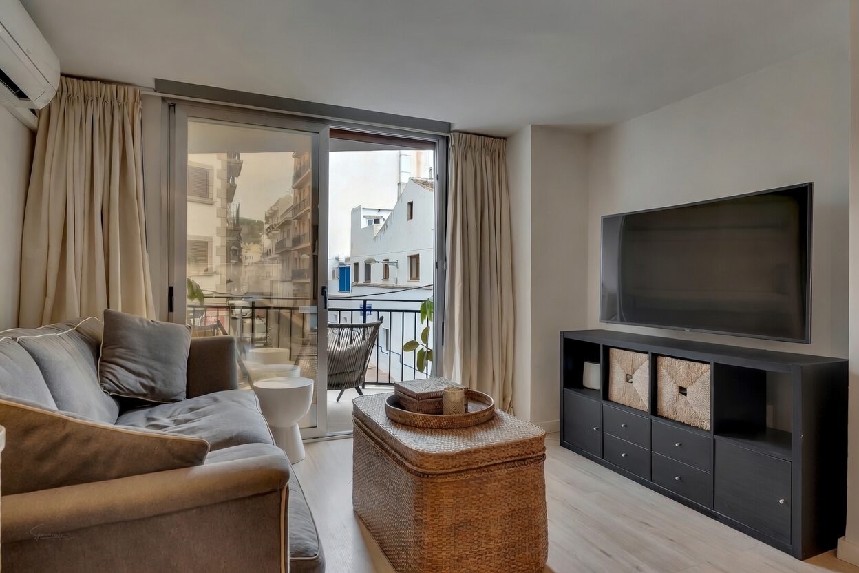 Salón luminoso con sofá gris, mesa de centro de mimbre y acceso a balcón con vistas urbanas.