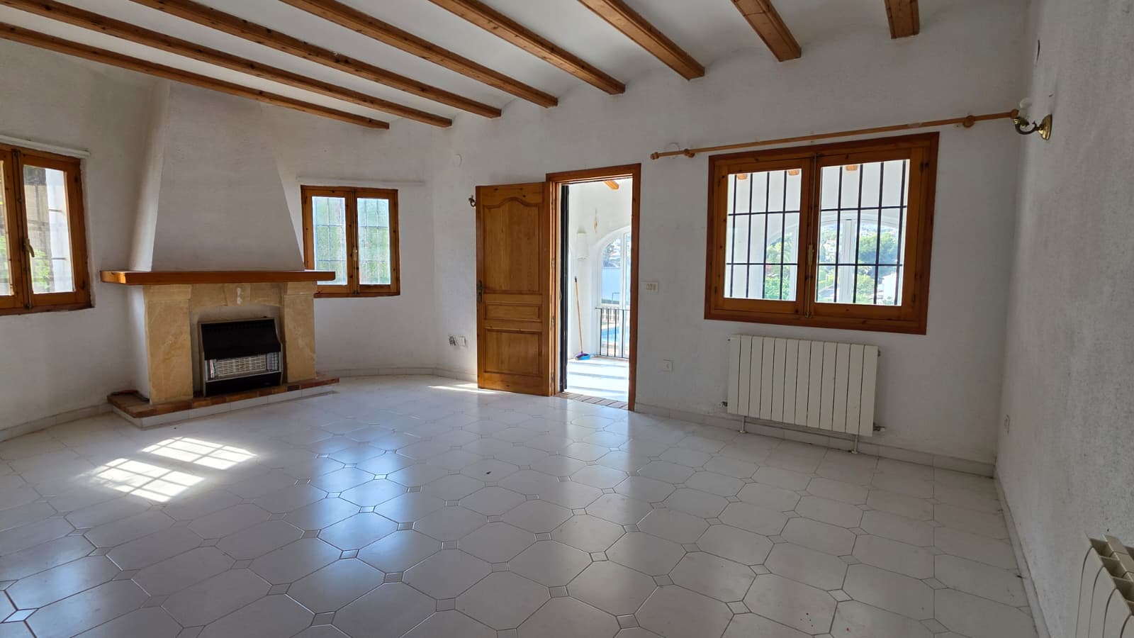 Salón con vigas de madera, chimenea de piedra tosca y suelo cerámico blanco. Ventanales de madera con acceso directo a la naya y luz natural.