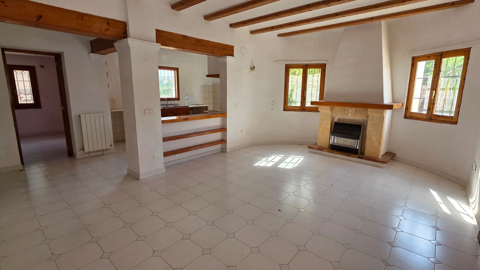 Salón espacioso con vigas de madera a la vista, chimenea de piedra con estufa integrada y suelos cerámicos. El espacio conecta con la cocina mediante una barra abierta.