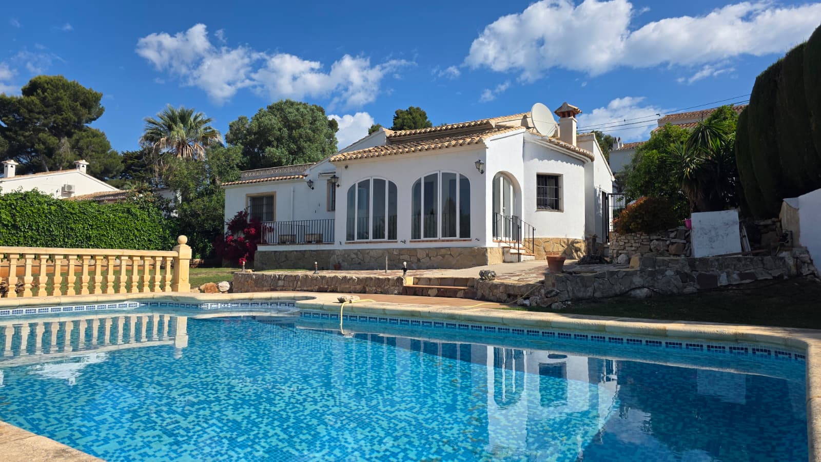 Vista exterior de la piscina privada y la fachada blanca con arcos acristalados y zócalo de piedra natural en la urbanización El Tosalet, Jávea.