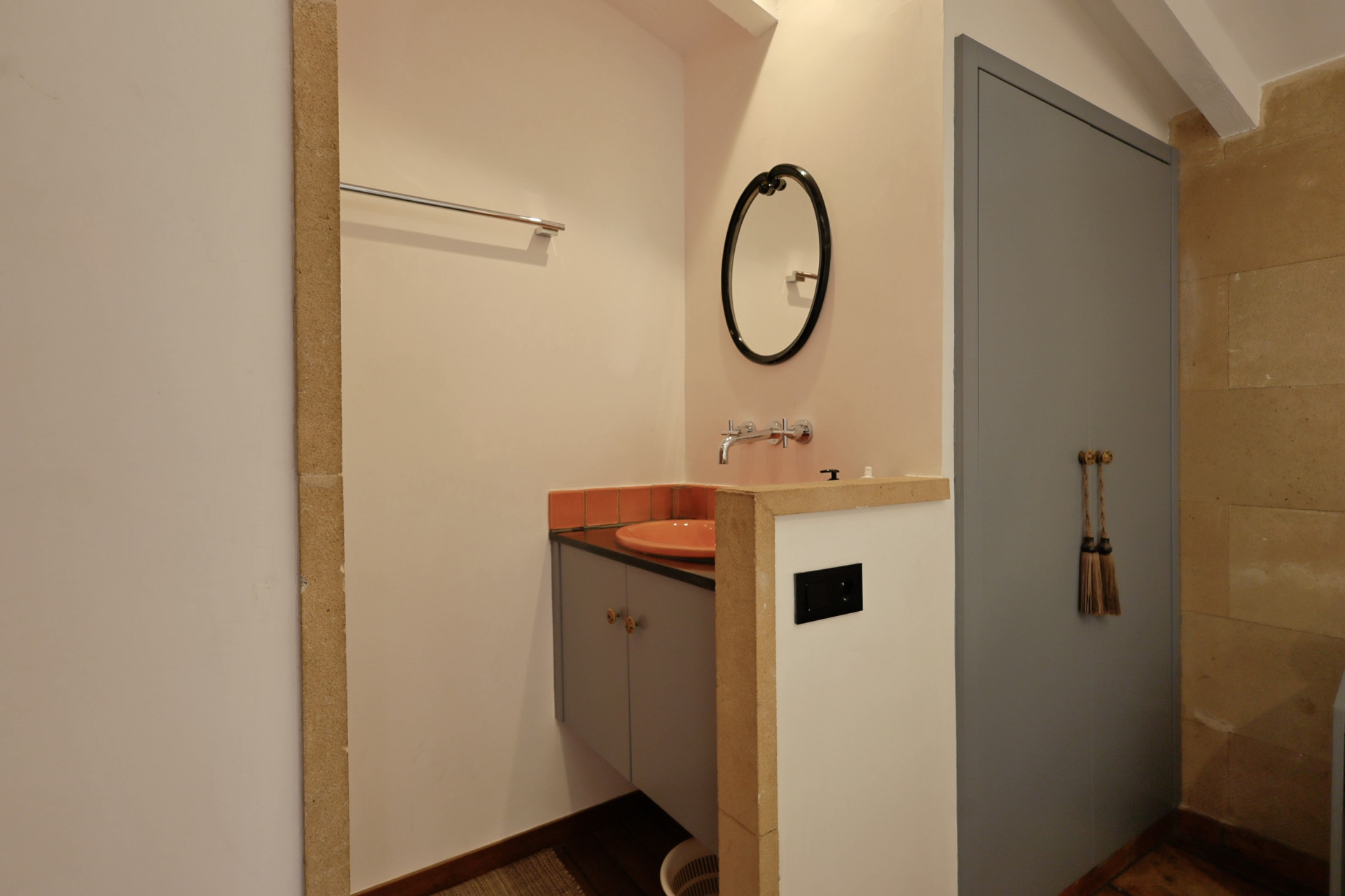 Detalle de baño con lavabo cerámico naranja, espejo ovalado y puerta gris con borlas decorativas.