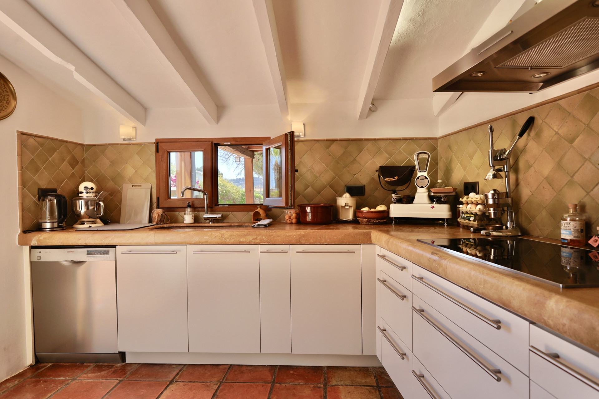 Detalle de la cocina con encimera de piedra y azulejos rústicos. Ventana de madera con vistas al exterior.