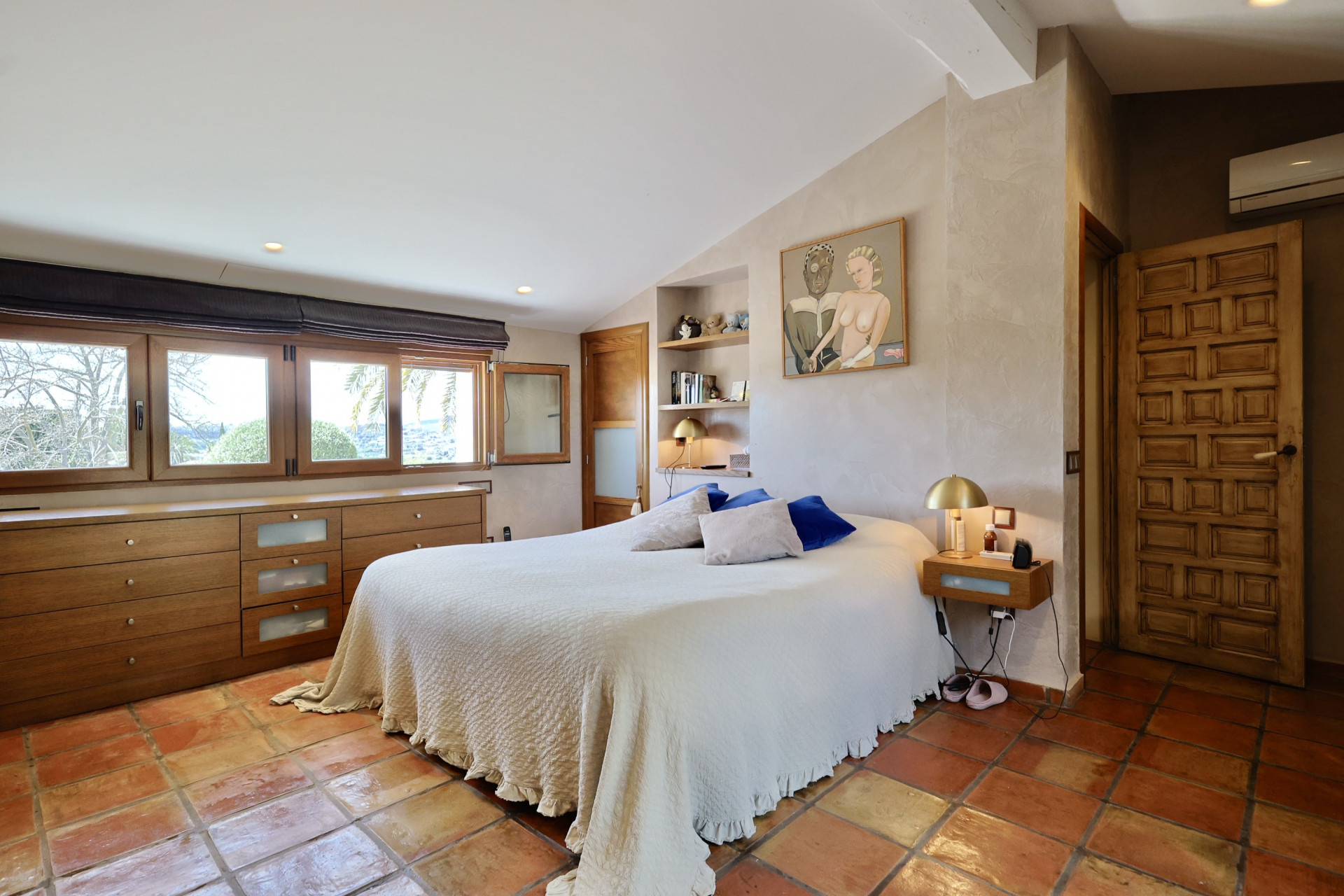 Dormitorio con suelo de terracota, cama doble con ropa de cama blanca y cabecera de madera con ventanas.