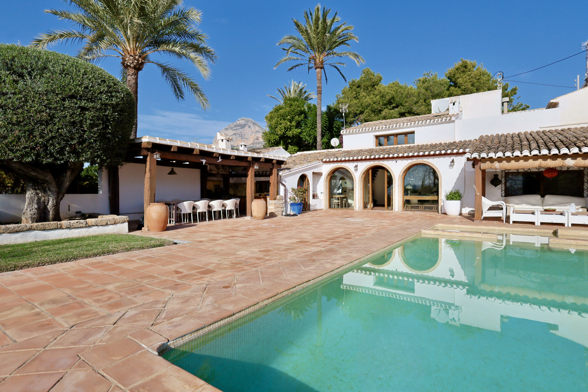 Vista de la piscina con baldosas de terracota, la terraza cubierta con asientos y los arcos de la villa.
