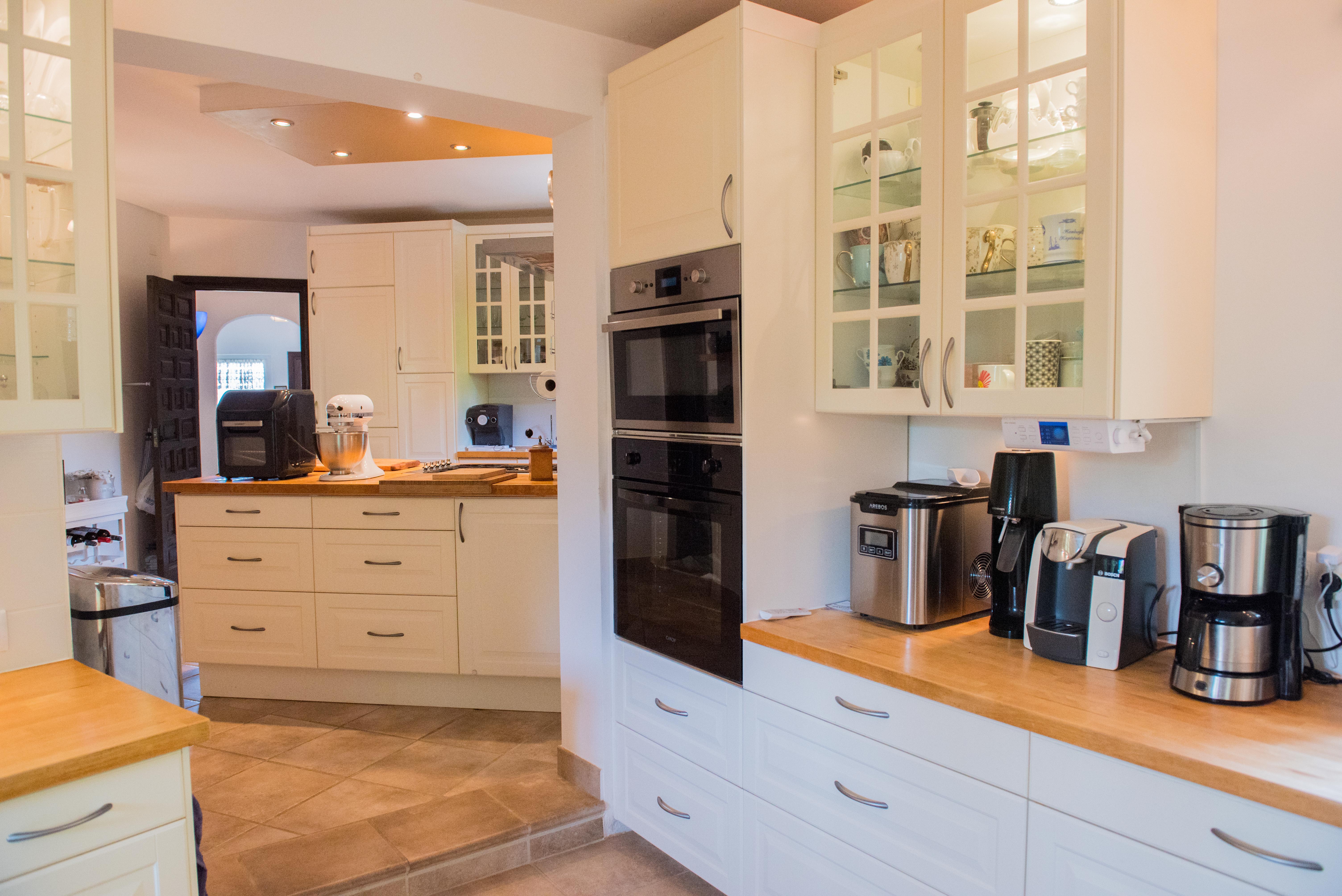 Cocina equipada con encimeras de madera natural, armarios color crema y hornos integrados. Dispone de vitrinas superiores, suelos cerámicos y abundante espacio de almacenamiento.