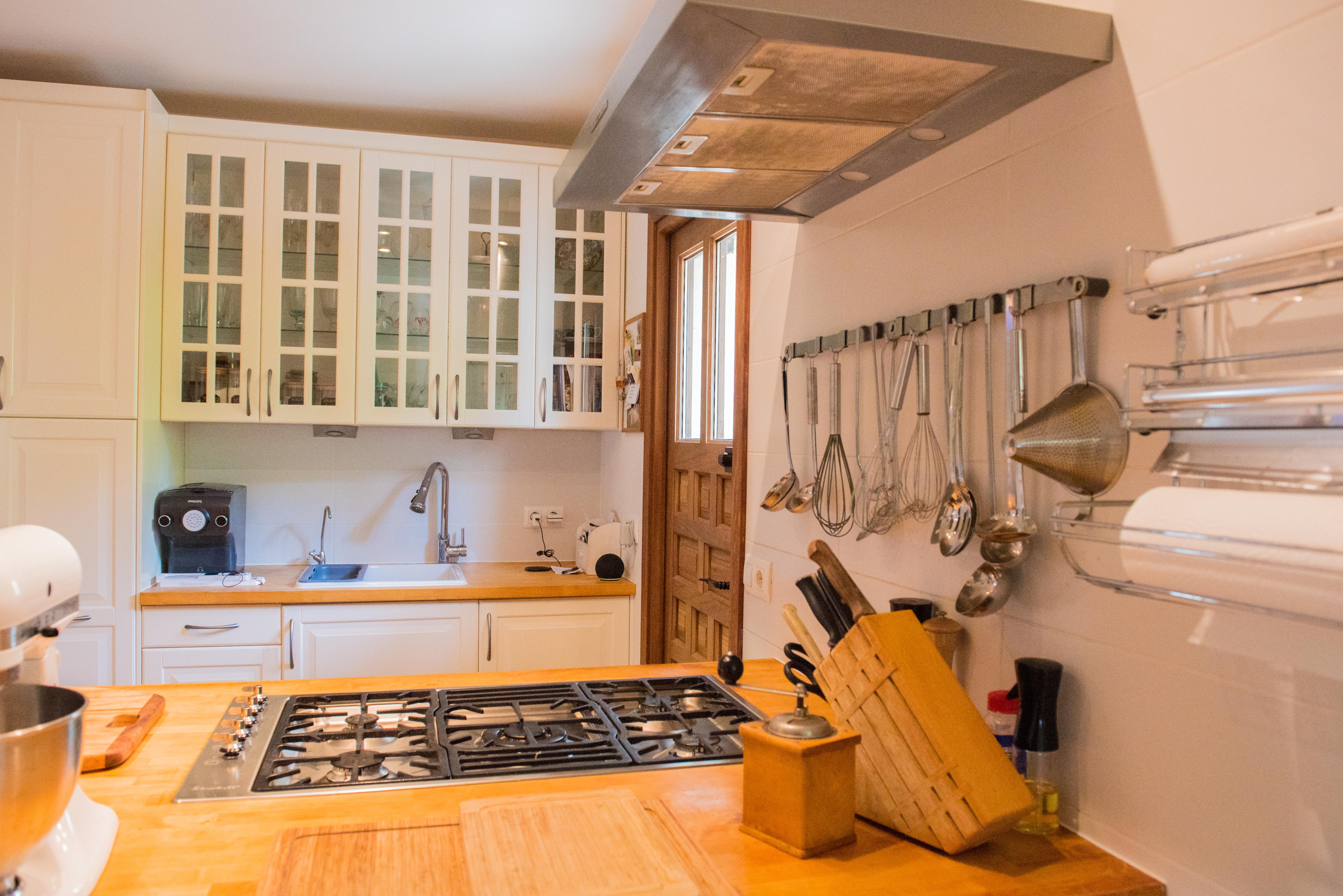 Cocina funcional con encimeras de madera, placa de gas integrada y armarios blancos con vitrinas superiores. Incluye zona de preparación con utensilios a la vista y puerta de madera.