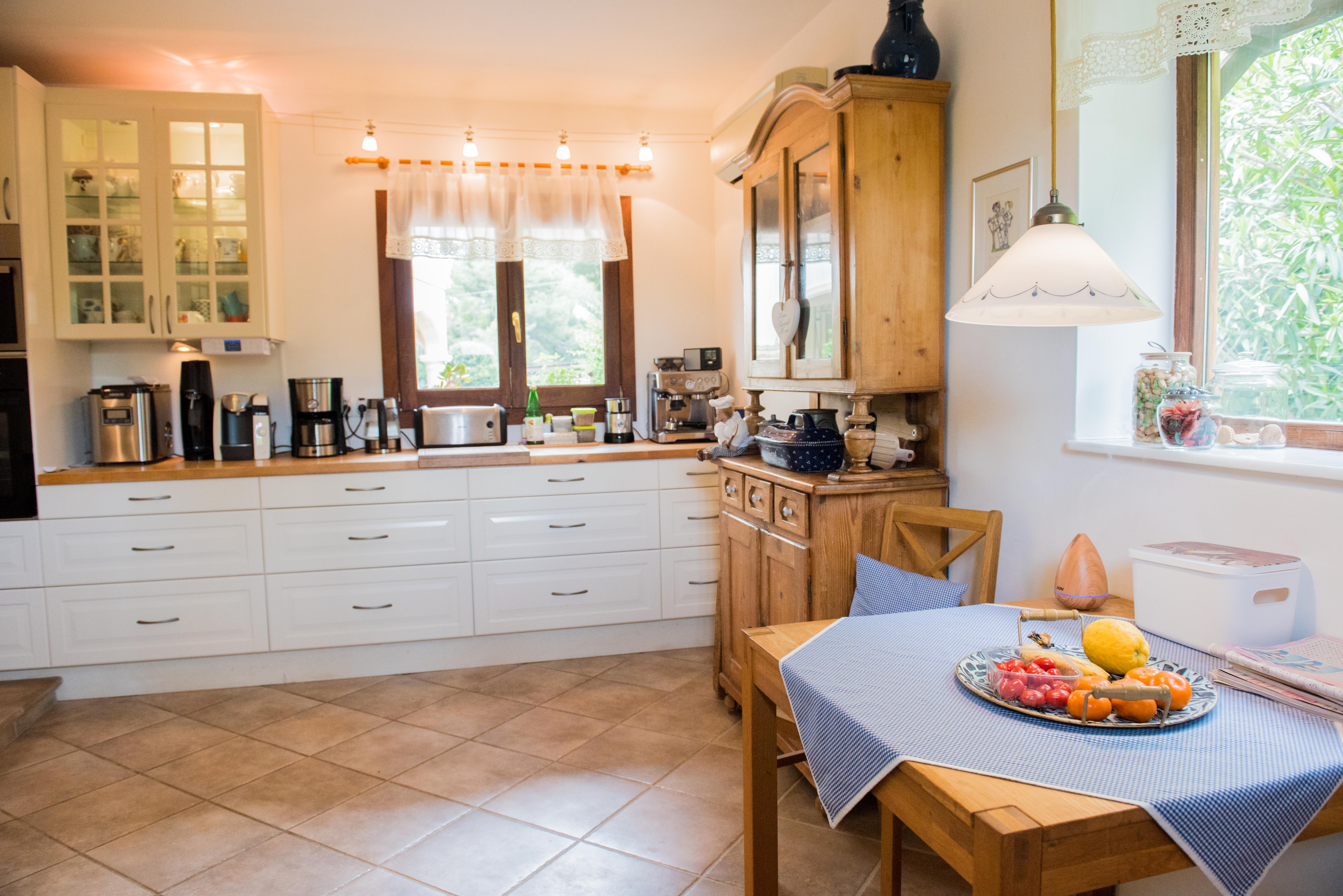 Cocina rústica con armarios blancos, encimeras de madera y zona de comedor. Incluye una alacena de madera clásica, suelo cerámico y luz natural desde dos ventanas.