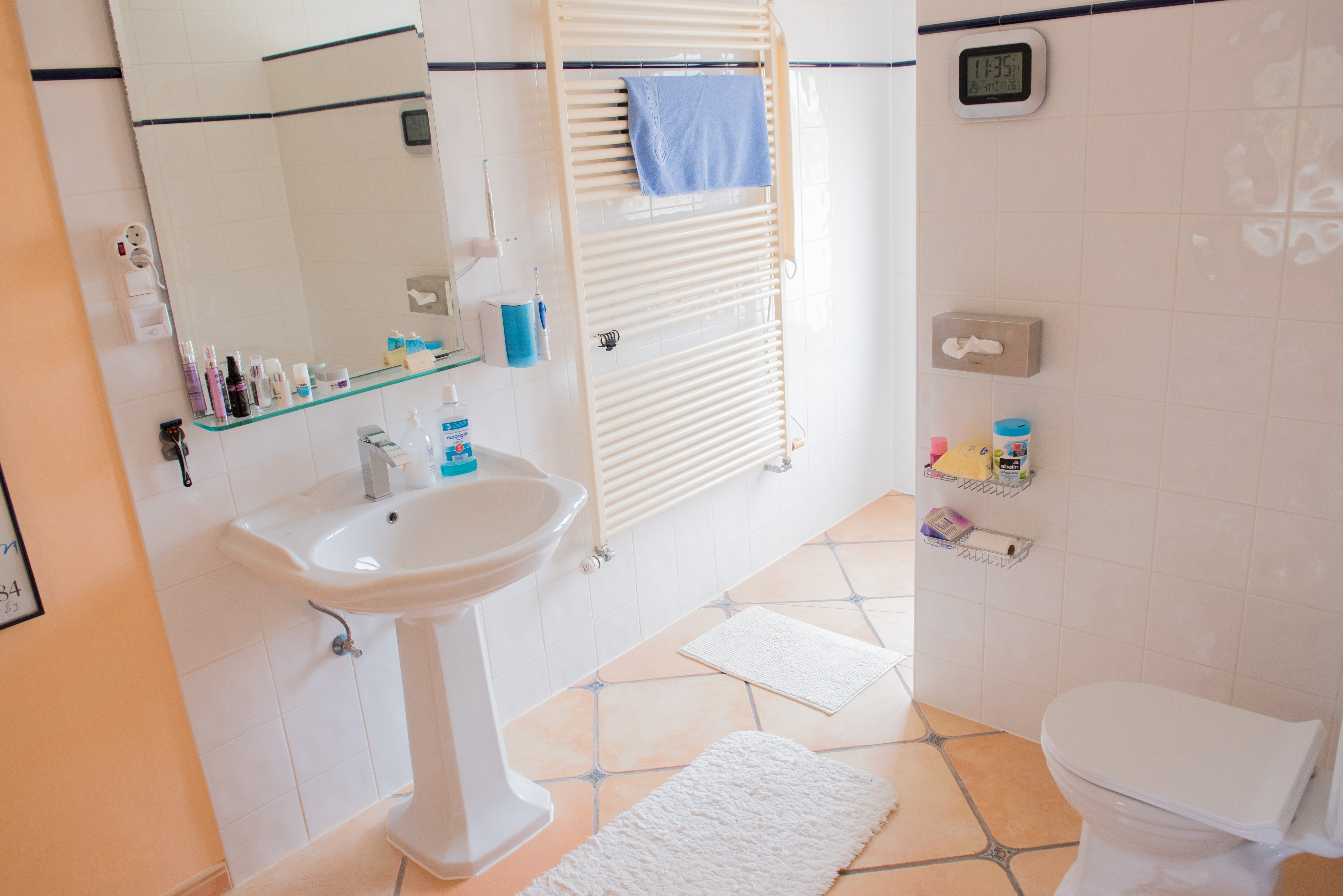 Cuarto de baño con lavabo de pedestal, suelos de terracota con detalles decorativos, radiador toallero y paredes alicatadas en blanco con cenefa azul.