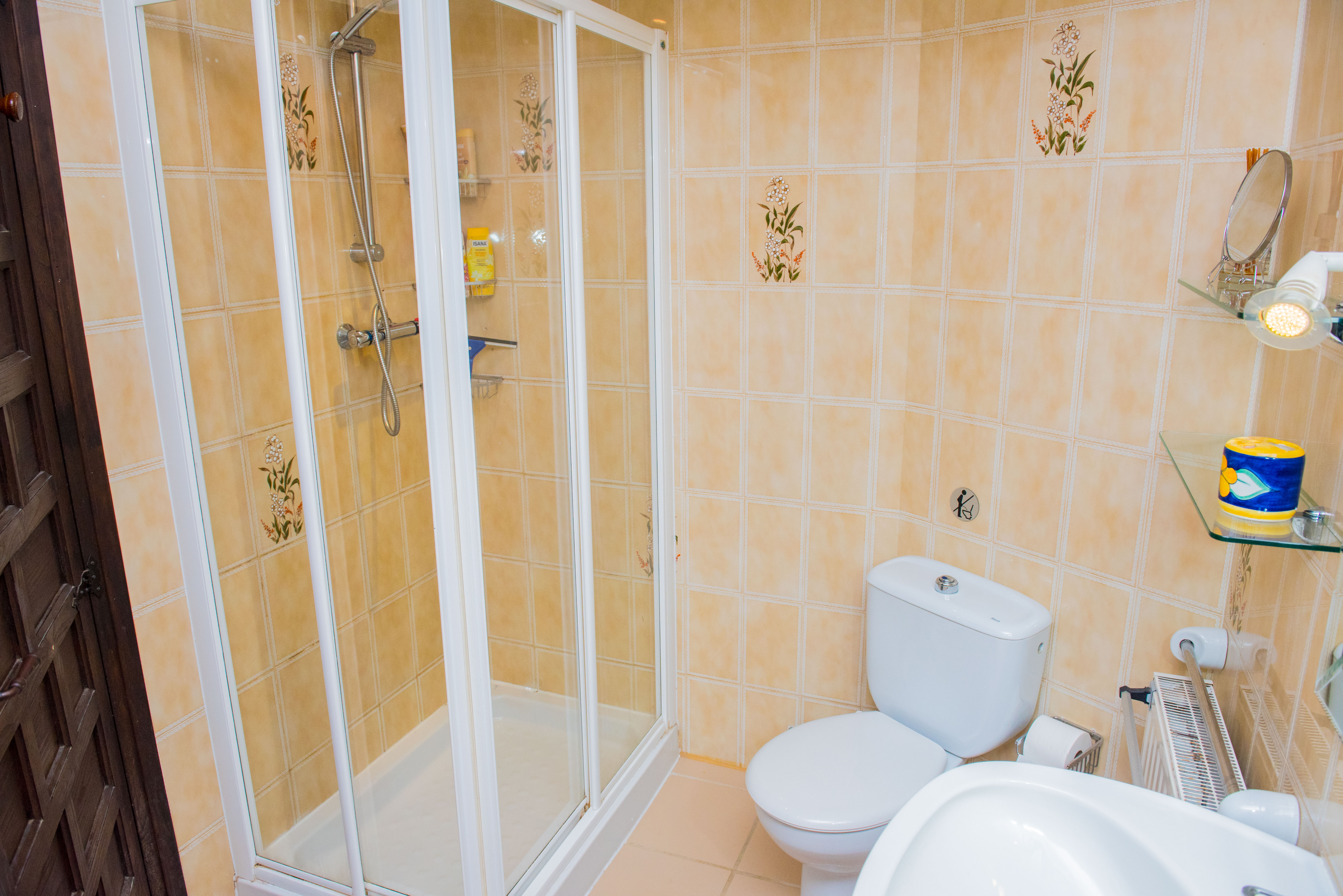 Cuarto de baño con mampara de ducha de marco blanco, paredes alicatadas en tonos beige con detalles florales y sanitarios de cerámica blanca.