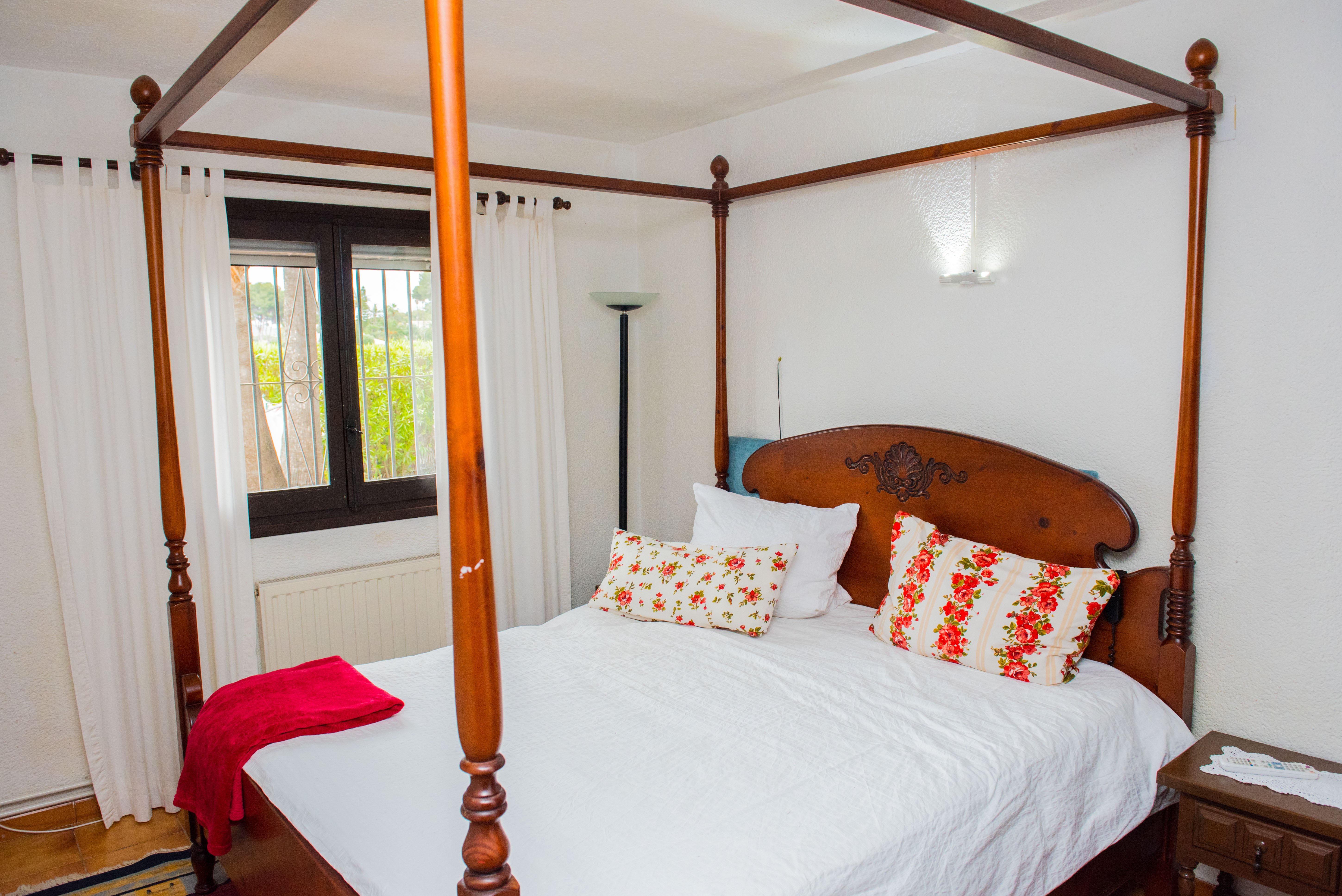 Dormitorio con cama de dosel de madera tallada, textiles florales y ventana con vistas al jardín. Incluye calefacción por radiador y paredes blancas.